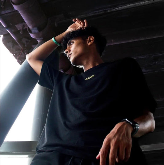 Young man in black Clover T-shirt posing under industrial ceiling with wristwatch and bracelet