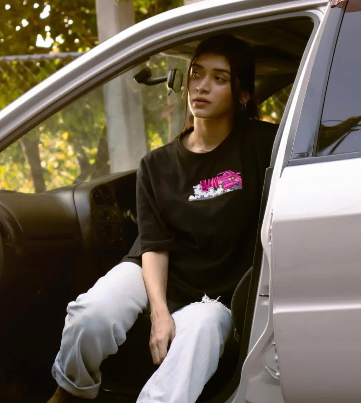 Young woman with black graphic t-shirt and ripped light jeans sitting in a car with open door during sunset