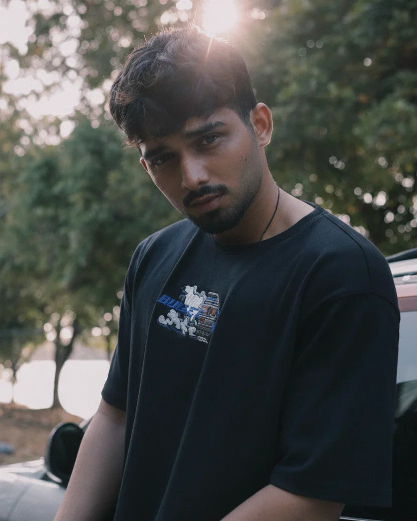 Young man with dark hair and beard wearing black t-shirt outdoors with trees and sunlight in background